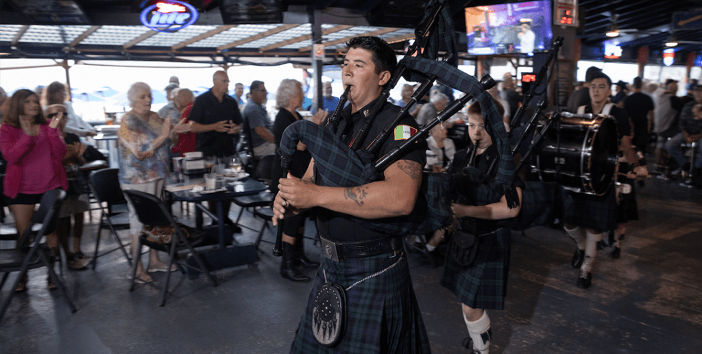 A man in black kilt playing bagpipes at an event.
